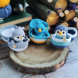 Könnte beinhalten: Drei gehäkelte Vogelornamente, jedes in einem gehäkelten eiförmigen Behälter. Die Vögel sind blau, grau und grau mit einem blauen Schnabel. Die Behälter sind weiß, gelb und blau.