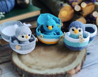 May include: Three crocheted bird ornaments, each in a crocheted egg-shaped container. The birds are blue, gray, and gray with a blue beak. The containers are white, yellow, and blue.