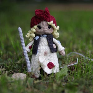 May include: A crocheted doll with blonde hair, wearing a red and white hat, a white dress with red and blue polka dots, and a black vest. The doll is holding a stick in its right hand.