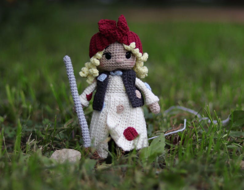 May include: A crocheted doll with blonde hair, wearing a red and white hat, a white dress with red and blue polka dots, and a black vest. The doll is holding a stick in its right hand.