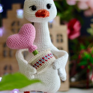 May include: A white crocheted goose holding a pink heart-shaped flower in a small pot with red and blue accents.