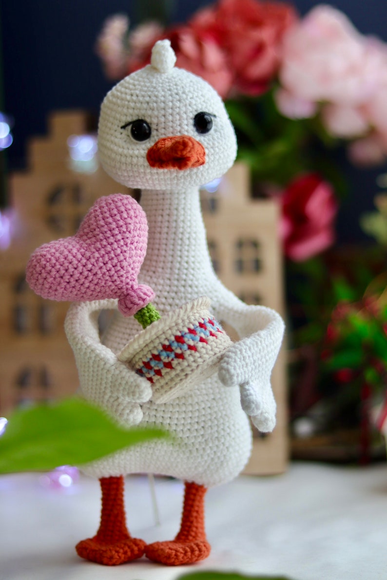 May include: A white crocheted goose holding a pink heart-shaped flower in a small pot with red and blue accents.