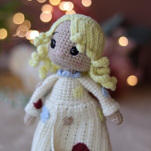 May include: A crocheted doll with blonde yarn hair, wearing a white dress with colorful patches. The doll has brown eyes and a sweet expression.