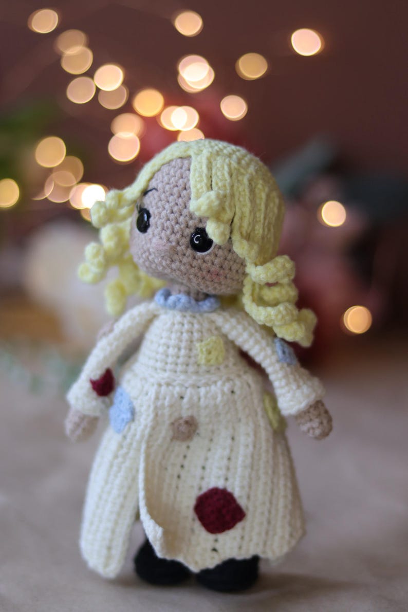 May include: A crocheted doll with blonde yarn hair, wearing a white dress with colorful patches. The doll has brown eyes and a sweet expression.