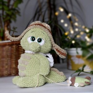 May include: A green crocheted plush toy with large eyes and a brown crocheted hat. The toy is holding a white crocheted scarf and is sitting on a white surface. A smaller green crocheted toy is lying on the surface.