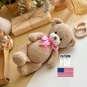 May include: Crochet pattern for a beige teddy bear with a pink bow. The pattern is available as a PDF download. The image also includes a US flag.