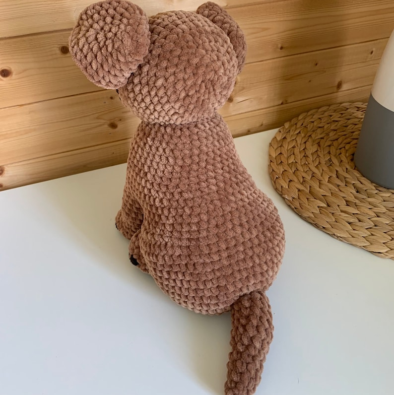 May include: A brown crocheted dog plush toy with a long tail. The dog is sitting on a white surface.