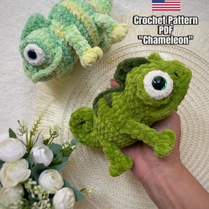 以下が含まれることがあります： 緑と黄色の2匹のぬいぐるみかぎ針編みカメレオン。1匹は横たわっており、もう1匹は手に持たれています。カメレオンは大きくて丸い目をしています。画像内のテキストは、アメリカ国旗とともに「Crochet Pattern PDF 'Chameleon'」と書かれています。