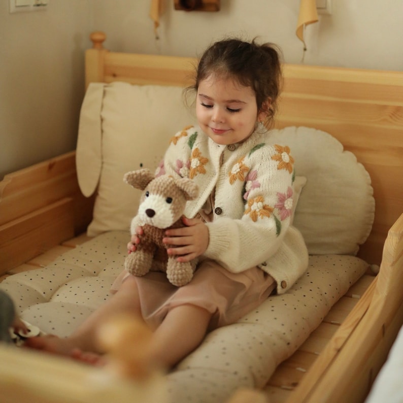 May include: A young girl wearing a white cardigan with floral embroidery sits on a bed holding a crocheted stuffed animal dog. The dog is brown and white with black eyes and a pink nose.