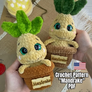 以下が含まれることがあります： 緑の葉とベージュの体を持つ2つの手編みのマンドレイク人形。それぞれ「マンドレイク」と書かれた茶色のポットに入っています。人形は青い目を持ち、ジュートの表面に展示されています。「Crochet Pattern 'Mandrake' PDF」というテキストが表示されています。