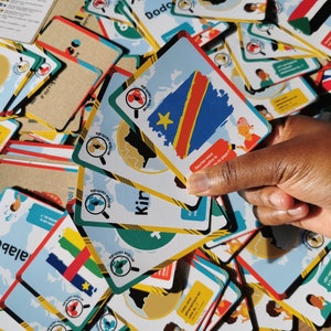 May include: A pile of flashcards with country flags and maps. The cards have a magnifying glass icon and French text. One hand is holding a card with the flag of the Democratic Republic of the Congo.