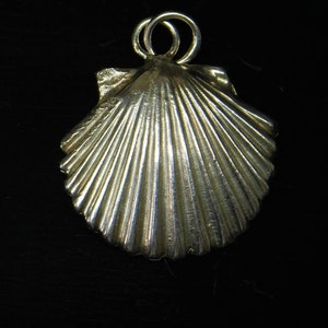 May include: A silver-colored seashell pendant with a loop for a chain, next to a 1956 quarter. The pendant has detailed ridges and a textured surface. The quarter is silver and shows the profile of George Washington.