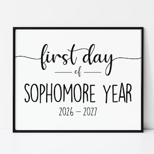 May include: A rectangular black frame containing a white sign with black text that reads first day of SOPHOMORE YEAR 2026 - 2027. The words first day are in a cursive script, while SOPHOMORE YEAR and the years are in a bold sans-serif font.