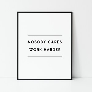 May include: A framed print with the text "NOBODY CARES WORK HARDER" in black, centered on a white background. The print is enclosed in a black frame, suitable for home or office decor.
