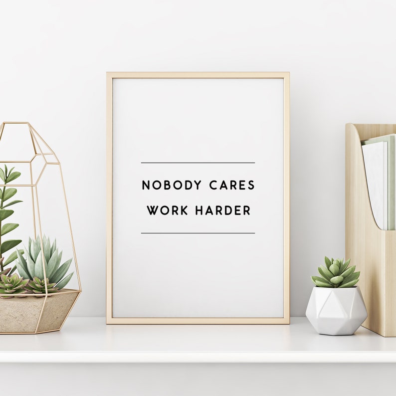 May include: A framed print with the text "NOBODY CARES WORK HARDER" in black, set against a white background. The frame is a light wood color. The print is displayed on a white shelf with decorative plants.