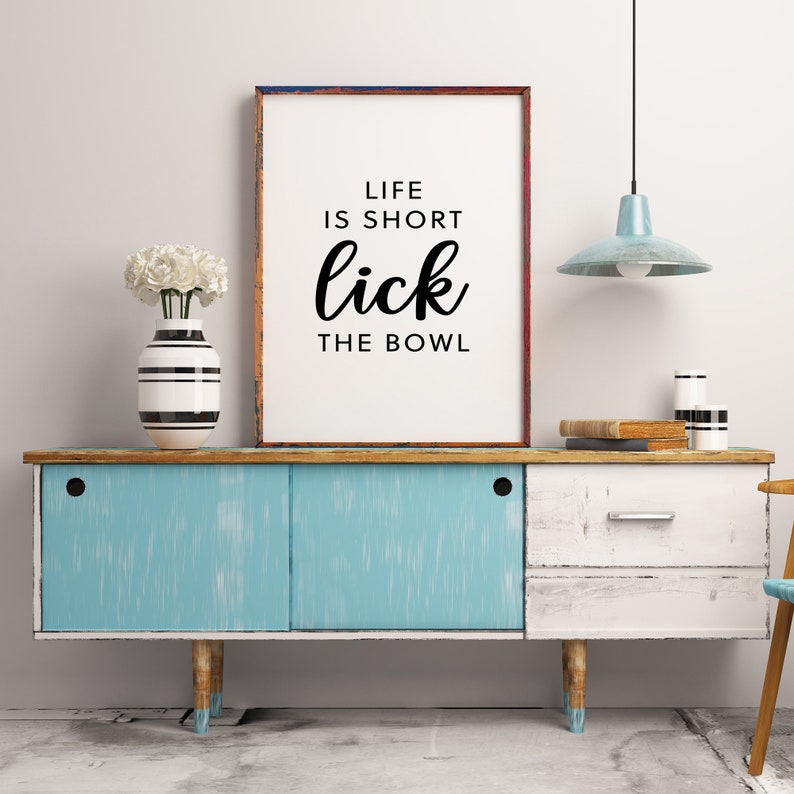 May include: A framed print with the text "LIFE IS SHORT lick THE BOWL" sits atop a distressed blue and white cabinet. A vase with white flowers and a striped vase are on the left. A light blue pendant lamp hangs above.