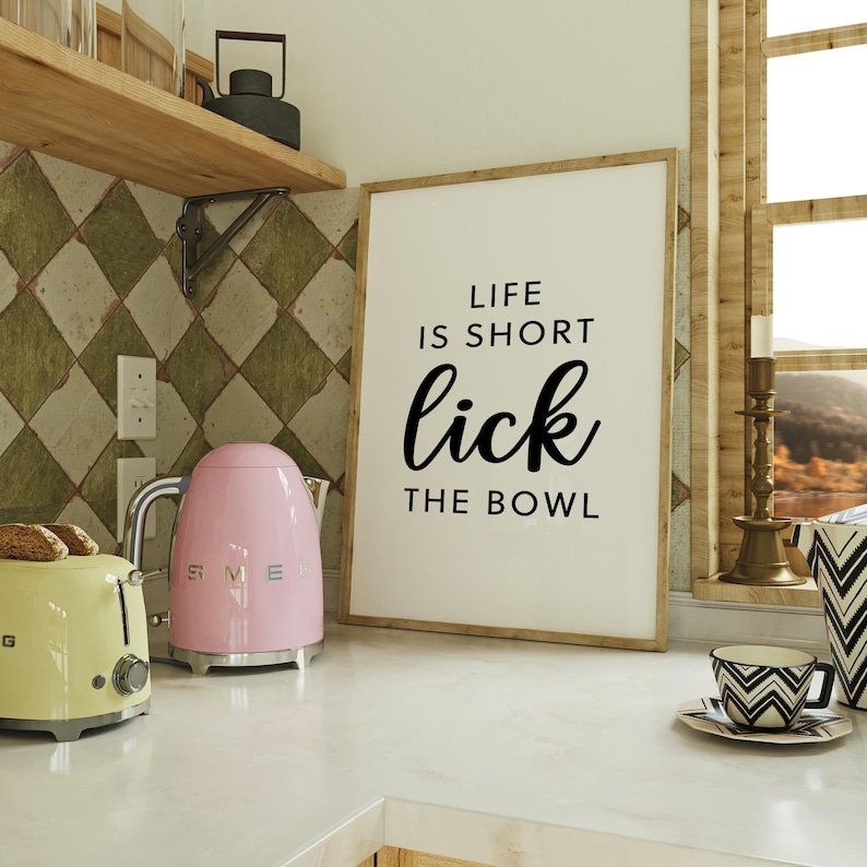 May include: A kitchen scene featuring a light pink SMEG kettle, a yellow toaster, and a framed print with the text "LIFE IS SHORT lick THE BOWL". The background includes a window, a patterned tile wall, and a decorative cup and saucer.