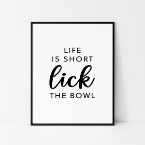 May include: A framed print with the text "LIFE IS SHORT lick THE BOWL" in black against a white background. The words are in a mix of fonts, with "lick" in a cursive style. The frame is black.