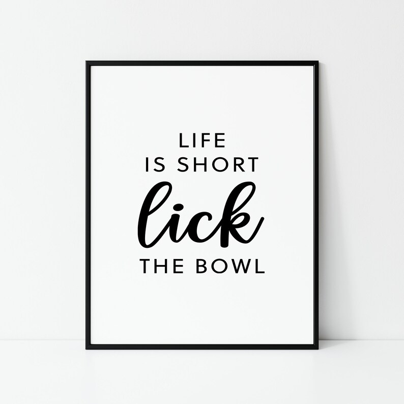 May include: A framed print with the text "LIFE IS SHORT lick THE BOWL" in black against a white background. The words are in a mix of fonts, with "lick" in a cursive style. The frame is black.