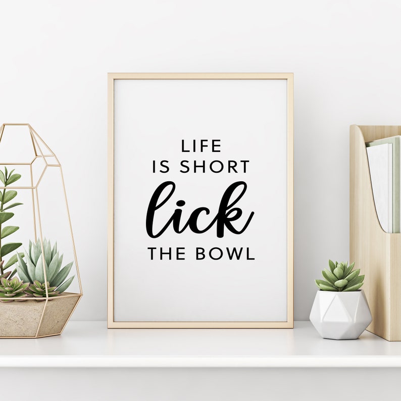 May include: A framed print with the text "LIFE IS SHORT lick THE BOWL" in black lettering. The frame is gold. The print is displayed on a white shelf with decorative plants and a wooden organizer.