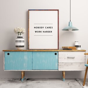 May include: A framed print with the text "NOBODY CARES WORK HARDER" sits atop a distressed blue and white cabinet. A vase with white flowers and a striped vase are on the cabinet. A light blue pendant lamp hangs above.