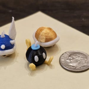 May include: Three small, 3D printed figurines of characters from the Super Mario Bros. video game series. The figurines are a blue Spiny Shell, a black Bob-omb, and a gold Koopa Troopa. The figurines are about the size of a US dime.