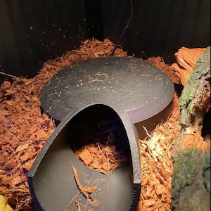 May include: Black plastic reptile hide with a curved tunnel entrance. The hide is sitting on a bed of brown wood chips.