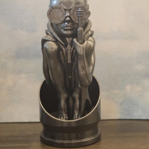 Könnte beinhalten: Eine silberfarbene Figur eines Menschen mit einer Brille und einer Kapuzenjacke. Die Figur steht auf einem zylindrischen Sockel.