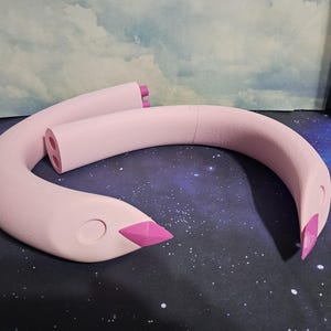 Può includere: Una scultura rosa chiaro e curva che ricorda il collo e la testa di un cigno. Il becco è di un rosa più scuro e la scultura ha una finitura liscia e opaca. La scultura del cigno è posta su uno sfondo stellato.