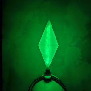 May include: A green light-up headband with a diamond-shaped light in the center. The light is on and illuminating the headband.