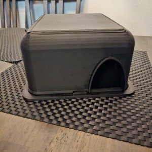 May include: A black plastic box with a rounded top and a small opening on one side. The box is sitting on a black woven placemat.