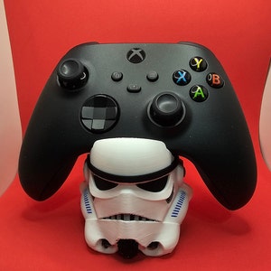 May include: A black Xbox controller is sitting on a white Stormtrooper figurine stand. The controller has a black and white checkered pattern on the back.