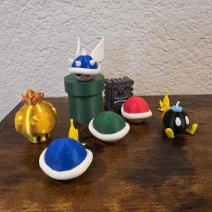 May include: A collection of 3D printed figures including a gold crown-topped mushroom, a blue winged shell on a green pipe, a grey stone block with a face, a red shell, a green shell, a blue shell, and a black bomb character with yellow feet.