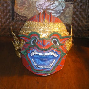 May include: A red, blue, and gold painted mask with a fierce expression. The mask has a large, open mouth with sharp teeth and a prominent nose. The eyes are wide and staring. The mask is decorated with gold trim and intricate details.