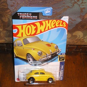 May include: Yellow Hot Wheels die-cast car model of a Volkswagen Beetle with a Transformers logo on the side. The car is on a blue and white card with the words "Hot Wheels" and "Transformers" printed on it.