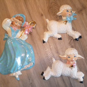 Puede incluir: Decoración de pared de cerámica vintage con una niña con un vestido azul y dos corderos blancos. La niña sostiene un cayado dorado y una flor rosa. Los corderos tienen lazos azules y rosas y pezuñas negras. Una adición encantadora.