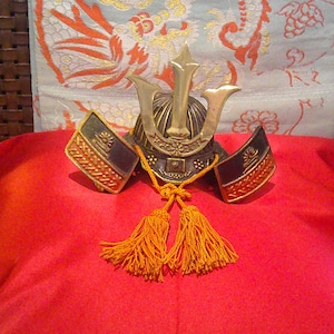 May include: A miniature Japanese samurai helmet, known as a kabuto, featuring a dark metallic bowl, a prominent gold-colored crest, and two side flaps. The helmet is adorned with a decorative orange cord tied in a knot with two hanging tassels.