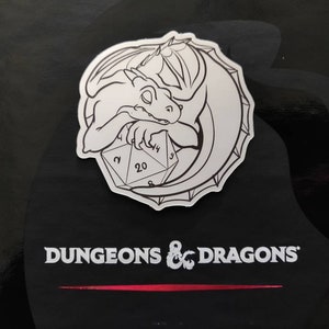 Op de afbeelding: Een zwart-witte sticker met een cartoon-draak die om een ​​twintigzijdige dobbelsteen is gekruld. De sticker heeft een witte achtergrond en een zwarte omtrek. De tekst "Dungeons & Dragons" is onder de sticker gedrukt.
