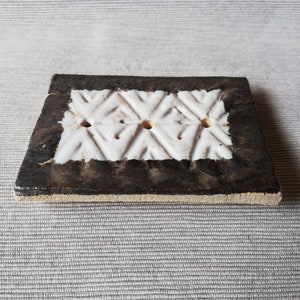 Op de afbeelding: Een witte keramische zeepbak met een geometrisch patroon en vier gaten. De bak heeft een zwarte rand en staat op een getextureerd oppervlak.