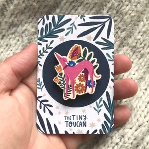 Op de afbeelding: Een roze houten pin van een hond met bloemmotieven, op een witte kaart met blauwe en groene bladeren en de tekst "The Tiny Toucan".