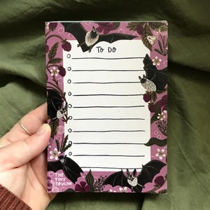 Op de afbeelding: Een rechthoekig notitieblok met een paarse rand met illustraties van vleermuizen, bloemen en bessen. Het notitieblok heeft een witte binnenkant met lijnen om op te schrijven en de woorden "TO DO" bovenaan. Het notitieblok wordt door een persoon vastgehouden.