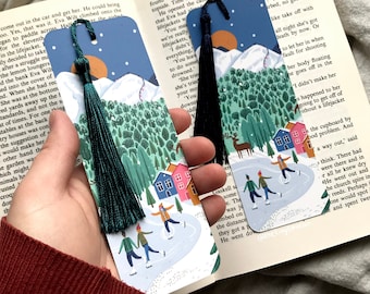 May include: Two colorful bookmarks with winter scenes. The bookmarks feature a snowy landscape with houses, trees, and people ice skating. One bookmark has a teal tassel and the other has a dark blue tassel.