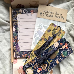 May include: A box containing stationery items with a witchy theme. Includes a notepad with a "To Do" list, two bookmarks with dark blue tassels, and a "Witches Brew Sticker Pack". The items feature illustrations of mushrooms, crystals, and potion bottles.