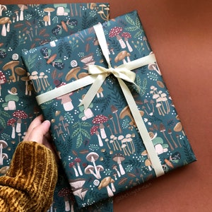 May include: Gift wrap featuring a whimsical mushroom and forest design in shades of teal, brown, and red. The paper is tied with a cream-colored ribbon. The pattern includes various mushroom illustrations, butterflies, and foliage.