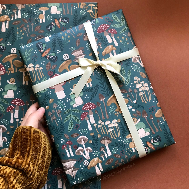 May include: Gift wrap featuring a whimsical mushroom and forest design in shades of teal, brown, and red. The paper is tied with a cream-colored ribbon. The pattern includes various mushroom illustrations, butterflies, and foliage.