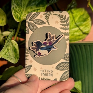 Op de afbeelding: Houten speld van een blauw-witte vogel met paarse bessen. De speld zit op een kaart met een grijze cirkel en een beige achtergrond met blad- en sterpatronen. De tekst "THE TINY TOUCAN" staat onderaan de kaart.