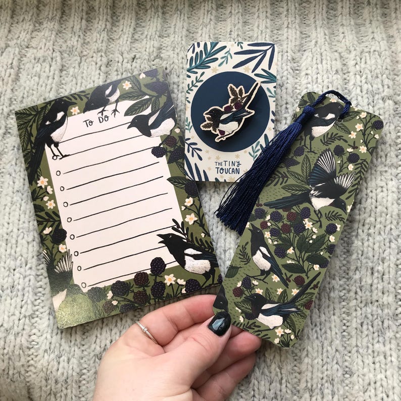 May include: A stationery set with a magpie design. The set includes a notepad with "To Do" text, a bookmark with a navy tassel, and a pin. The items feature a green background with magpies and floral illustrations.