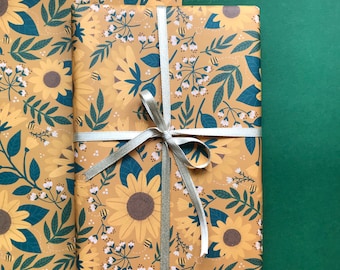 May include: A yellow and green floral gift wrap with a silver ribbon. The pattern features sunflowers, leaves, and small white flowers. The text "@thetinytoca" is printed in the top right corner.