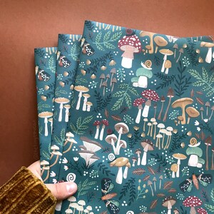 May include: Three folded fabric pieces with a teal background and a whimsical mushroom pattern. The design includes various mushroom types, leaves, butterflies, and snails in shades of brown, red, and white. The fabric is held by a person wearing a brown corduroy sleeve.
