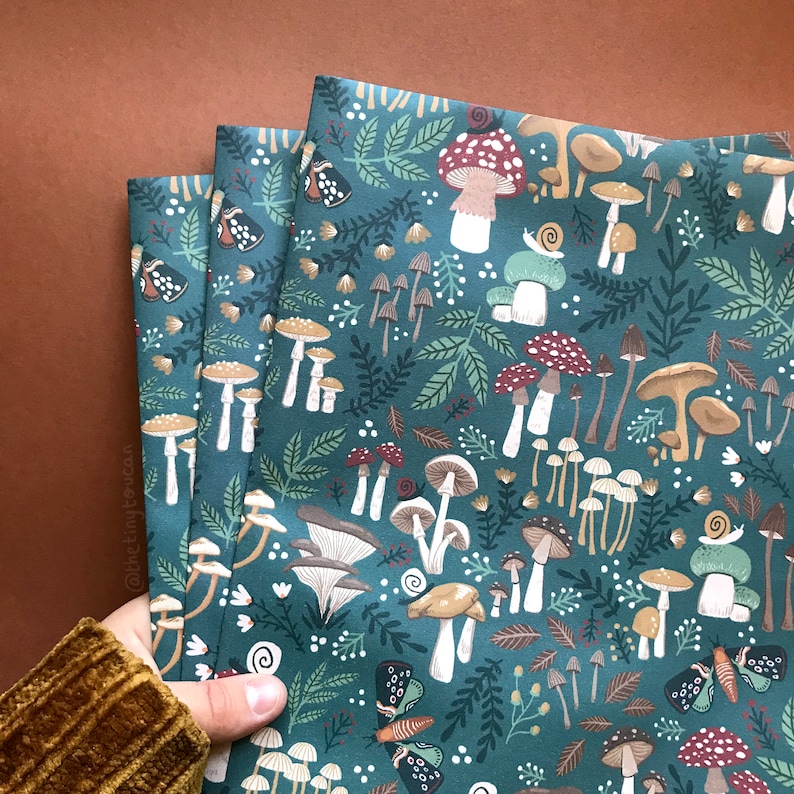 May include: Three folded fabric pieces with a teal background and a whimsical mushroom pattern. The design includes various mushroom types, leaves, butterflies, and snails in shades of brown, red, and white. The fabric is held by a person wearing a brown corduroy sleeve.